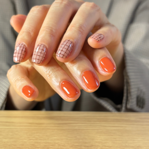 テラコッタのワンカラーとブラウンチェックのアシンメトリーネイルでポージングしている手元のアップ